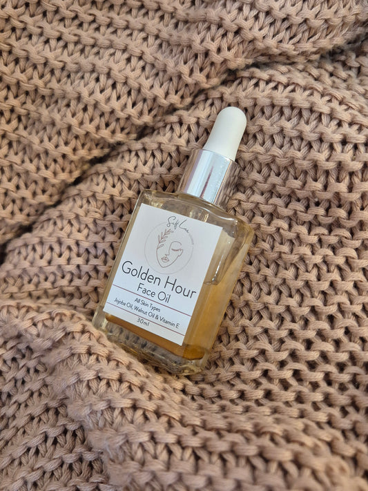A bottle of Golden Hour Face Oil by The Skin Collective, featuring a clear glass bottle with a white dropper cap and a white label, resting on a beige knitted blanket.