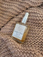 A bottle of Golden Hour Face Oil by The Skin Collective, featuring a clear glass bottle with a white dropper cap and a white label, resting on a beige knitted blanket.